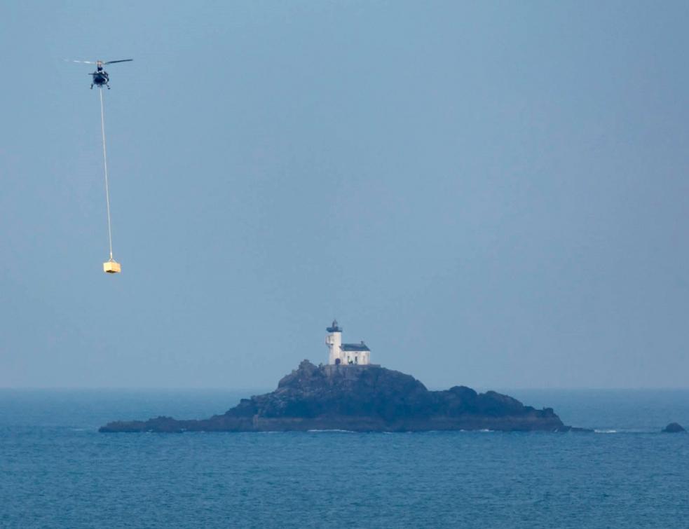 Le phare de Tévénnec au loin, un hélicoptère apporte du matériel le long d'une corde.